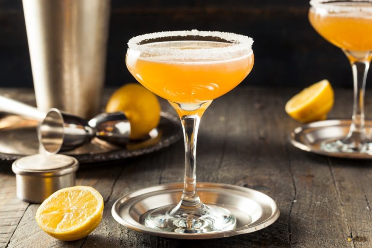 Classic sidecar cocktail with a sugar rim displayed with a moody background, oranges and a cocktail shaker
