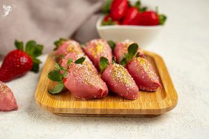 Pink chocolate-dipped strawberries with gold and white sprinkles arranged on a wooden tray for a party treat.