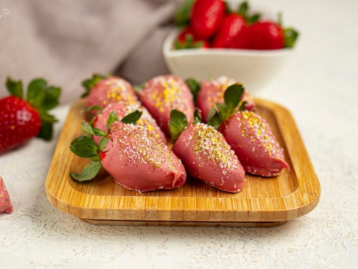 Pink chocolate-dipped strawberries with gold and white sprinkles arranged on a wooden tray for a party treat.