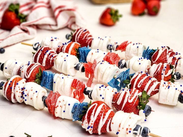 Red, white, and blue dessert skewers with strawberries, marshmallows, candy strips, blueberries, white chocolate drizzle and patriotic sprinkles.
