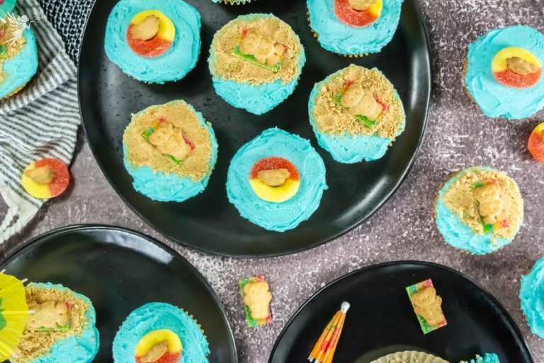 Beach-themed cupcakes with blue frosting, teddy graham bears, gummy rings, and crushed cookie sand on black serving plate.