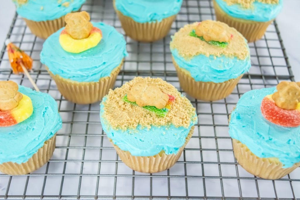 Beach-themed cupcakes with blue frosting, teddy graham bears, gummy rings, and crushed cookie sand on cooling rack.