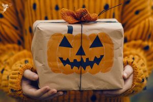 a child holding a brown paper package wrapped with orange bow and a pumpkin face, gifting for halloween