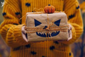 a lady in orange wool sweater carrying a brown paper package thats been decorated with a scary halloween face and wrapped in orange string