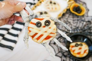 A hand holding a mini mummy pizza on a skeleton hand prop, showing the cheese strip bandages and olive eyes.