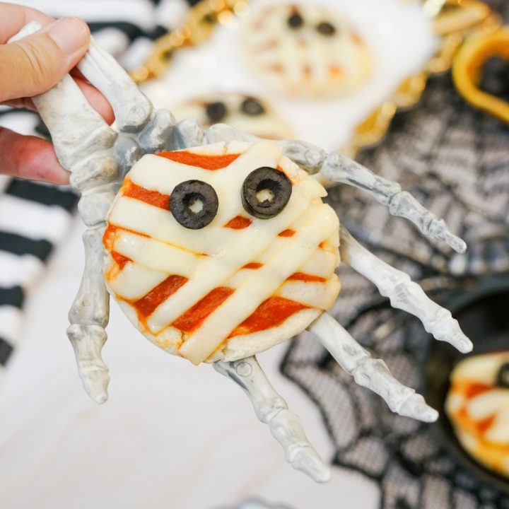 A hand holding a mini mummy pizza on a skeleton hand prop, showing the cheese strip bandages and olive eyes.