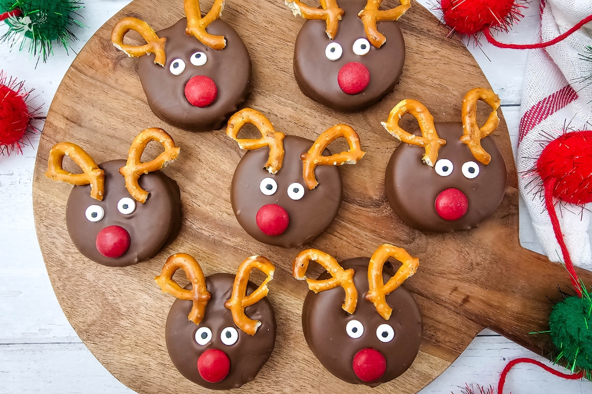 A group of seven adorable finished reindeer Oreo cookies arranged on a round wooden serving board with festive red and green pom-poms.