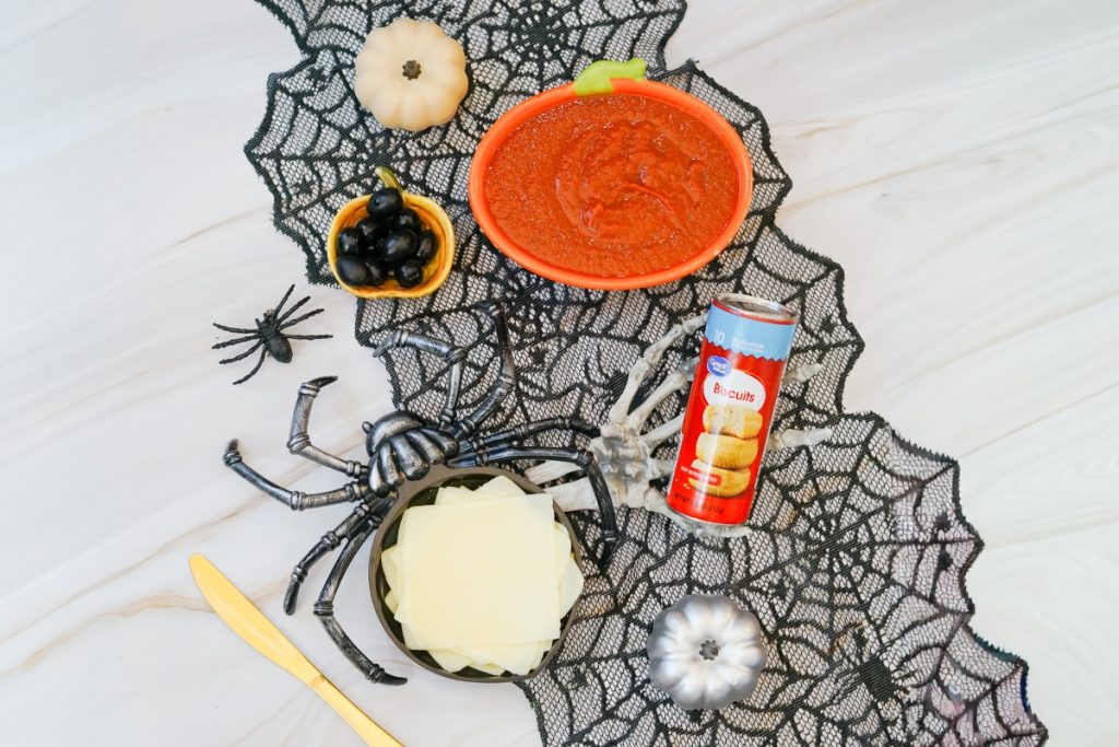 Mummy pizza ingredients—biscuits, sauce, cheese slices, and olives—arranged with Halloween spider decorations on a spiderweb runner.