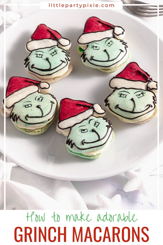Adorable Grinch macarons with green faces, red icing hats, and white filling, presented on a clean white platter.