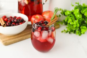 A refreshing glass of Sparkling Cranberry Apple Cider Punch with ice, cranberries, a cinnamon stick, and rosemary sprig, with a pitcher in the background.