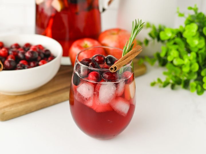 A refreshing glass of Sparkling Cranberry Apple Cider Punch with ice, cranberries, a cinnamon stick, and rosemary sprig, with a pitcher in the background.