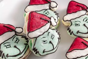 A close-up of several Grinch-themed macarons with green faces, red Santa hats, and dark chocolate piped features.