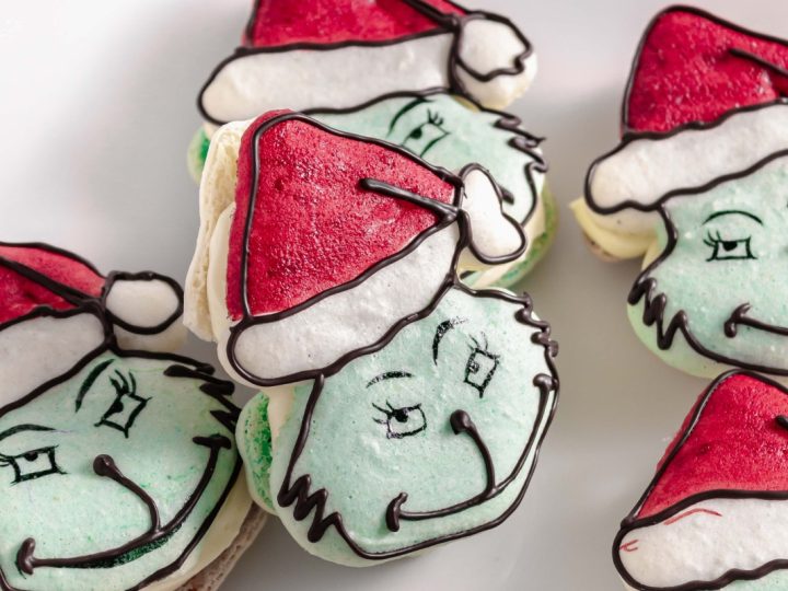 A close-up of several Grinch-themed macarons with green faces, red Santa hats, and dark chocolate piped features.