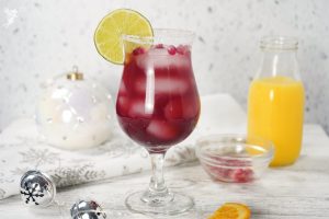 A vibrant red Pomegranate Margarita on a whitewashed table with Christmas ornaments and an orange juice bottle.