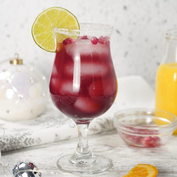 A vibrant red Pomegranate Margarita on a whitewashed table with Christmas ornaments and an orange juice bottle.