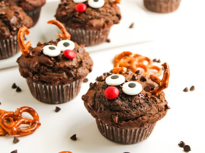 Two festive Reindeer Double Choc Chip Muffins with pretzel antlers, candy eyes, and a red nose, ready for a holiday party.