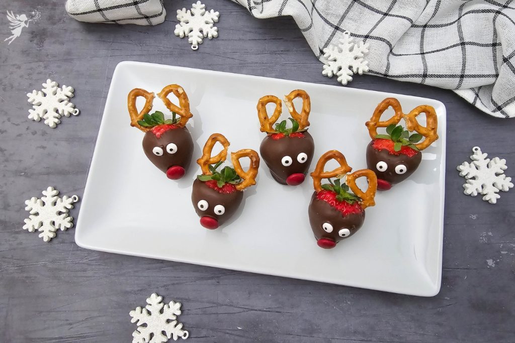 Five completed Reindeer Strawberries arranged on a white rectangular plate, surrounded by decorative white snowflakes.