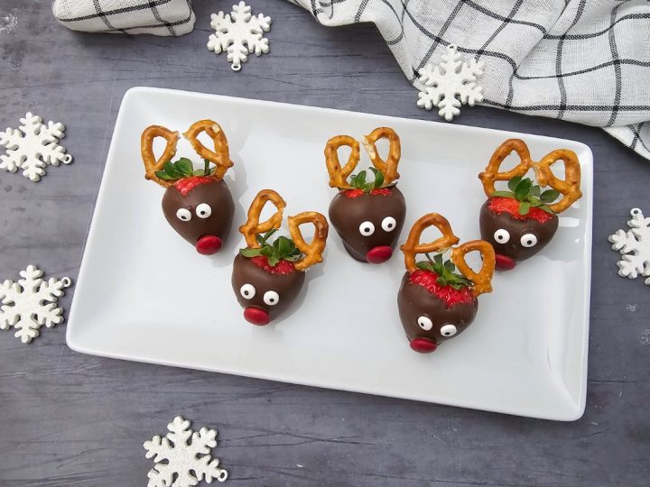 Five completed Reindeer Strawberries arranged on a white rectangular plate, surrounded by decorative white snowflakes.