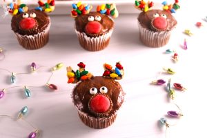 A group of four adorable reindeer-themed cupcakes on a white surface, surrounded by scattered miniature Christmas light candies.