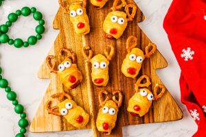 Baked Rudolph Reindeer Pigs in Blankets appetizers decorated with pretzel antlers, candy eyes and ketchup noses, placed on a festive cutting board.