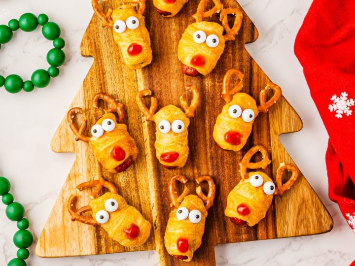 Baked Rudolph Reindeer Pigs in Blankets appetizers decorated with pretzel antlers, candy eyes and ketchup noses, placed on a festive cutting board.