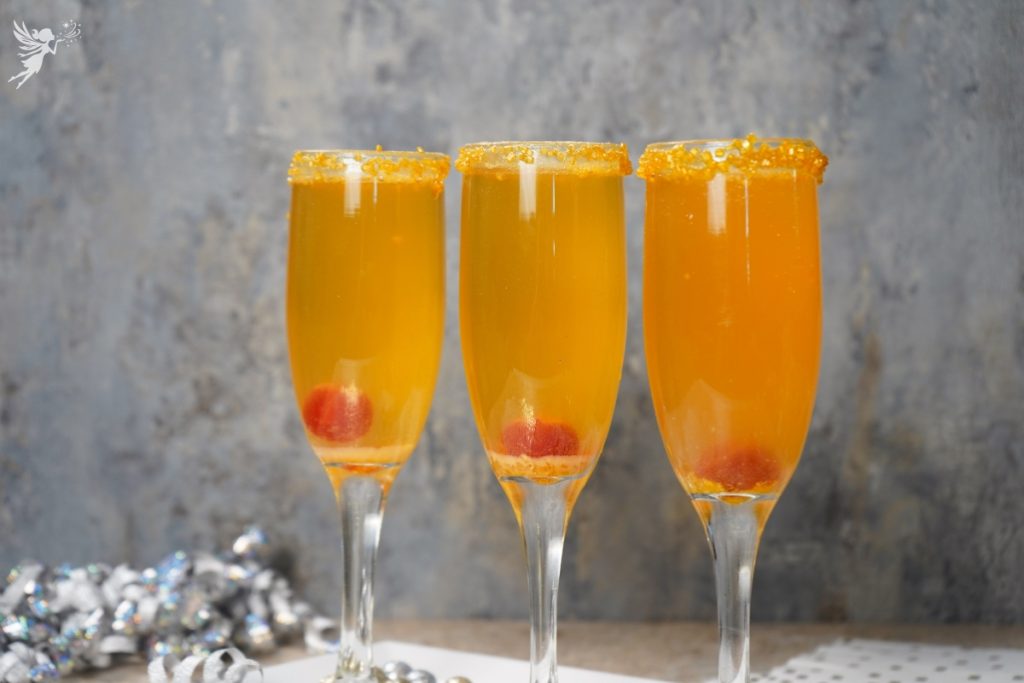 Three flutes of the vibrant Shimmering Golden Champagne Cocktail, garnished with a gold sugar rim and a cherry.