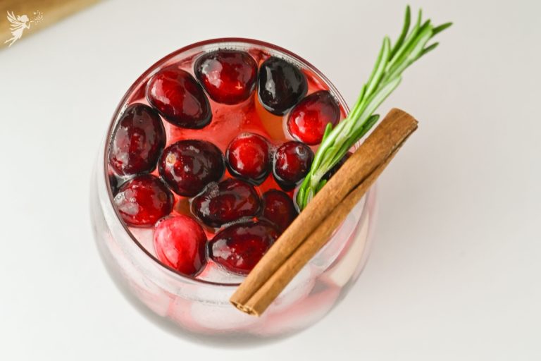 Close-up overhead shot of a glass filled with Sparkling Cranberry Apple Cider Punch, garnished with floating cranberries, a cinnamon stick, and fresh rosemary.