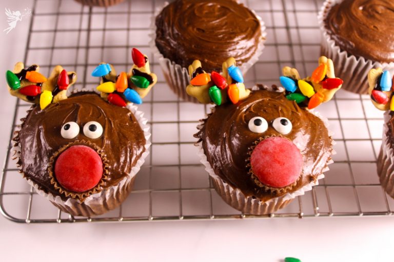 Two finished reindeer cupcakes cooling on a wire rack, complete with chocolate frosting, festive cookie antlers, red noses, and googly eyes.