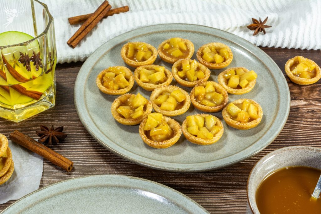 A plate of finished mini Caramel Apple Tartlets with diced apple filling, next to cinnamon sticks and star anise.