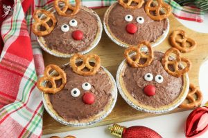 Four adorable Rudolph Mini Pies on a wooden board, featuring pretzel antlers and red candy noses for a festive treat.