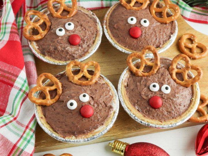 Four adorable Rudolph Mini Pies on a wooden board, featuring pretzel antlers and red candy noses for a festive treat.