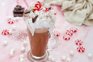 Full mug of Christmas Peppermint Hot Chocolate surrounded by peppermint candies and mini marshmallows on a pink surface.