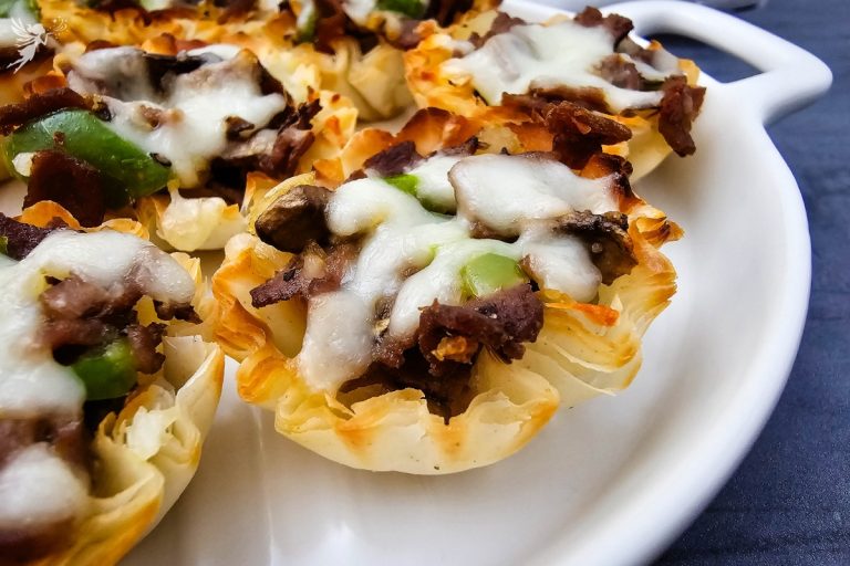 Close-up of baked Philly Cheesesteak Phyllo Bites on a white plate with melted white cheese and cooked green peppers.