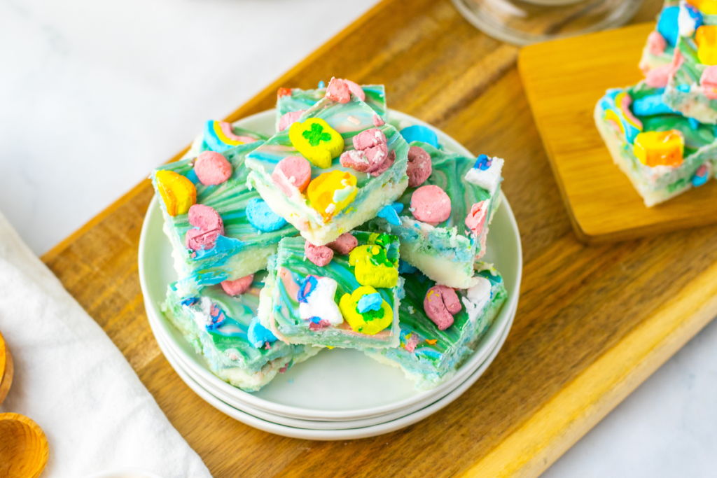 A stack of finished, square-cut pieces of marbled green, blue, and pink Lucky Charms Fudge, topped with colorful marshmallow shapes, on a white plate.