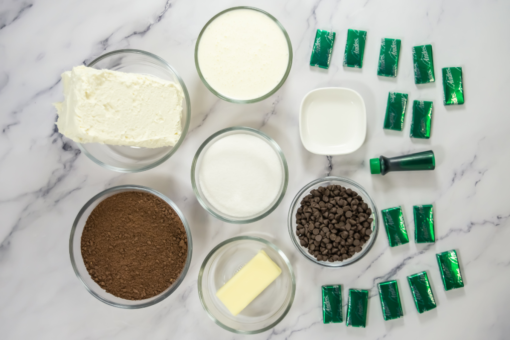 Overhead view of all ingredients for Mini Mint Cheesecakes: cream cheese, chocolate crumbs, Andes mints, chips, sugar, and green food coloring on marble.