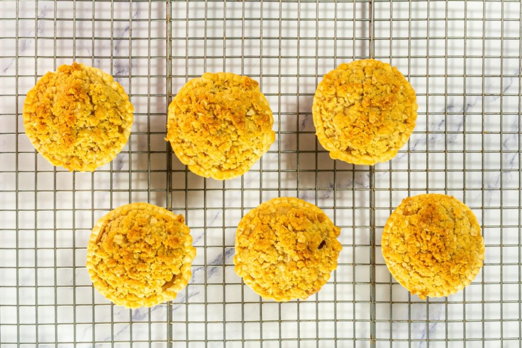 Six freshly baked Mini Dutch Apple Pies that is part of Mini Pie Dessert Platter are cooling on a wire rack, featuring a golden-brown, crumbly topping.