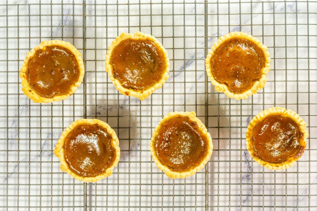 Six freshly baked Mini Pumpkin Pies that is part of Mini Pie Dessert Platter are cooling on a metal wire rack, featuring a rich, dark orange filling and golden crusts.