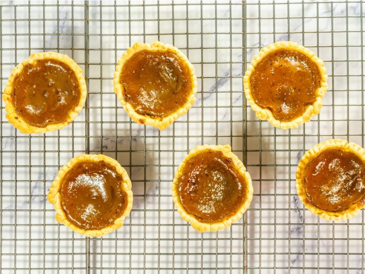 Six freshly baked Mini Pumpkin Pies that is part of Mini Pie Dessert Platter are cooling on a metal wire rack, featuring a rich, dark orange filling and golden crusts.