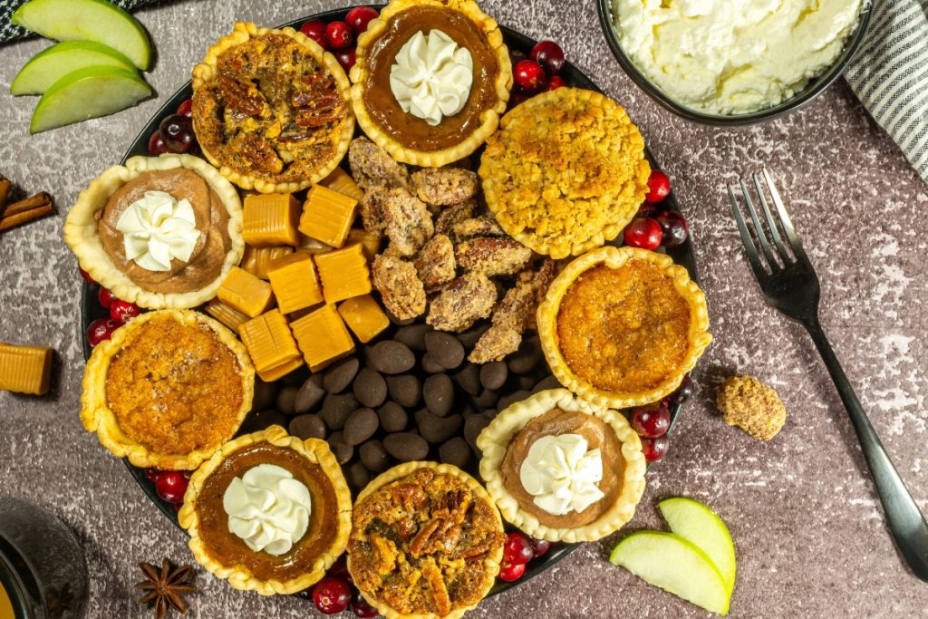 A beautifully arranged Mini Pie Dessert Platter featuring different mini pies decorated with whipped cream, surrounded by candies, chocolate drops, cranberries, and apple slices, ready for serving.