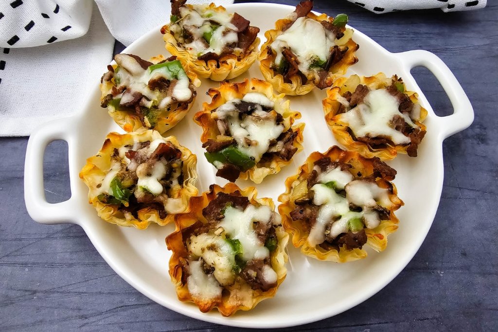 Final image of seven baked Philly Cheesesteak Phyllo Bites with melted cheese on a handled white serving dish.
