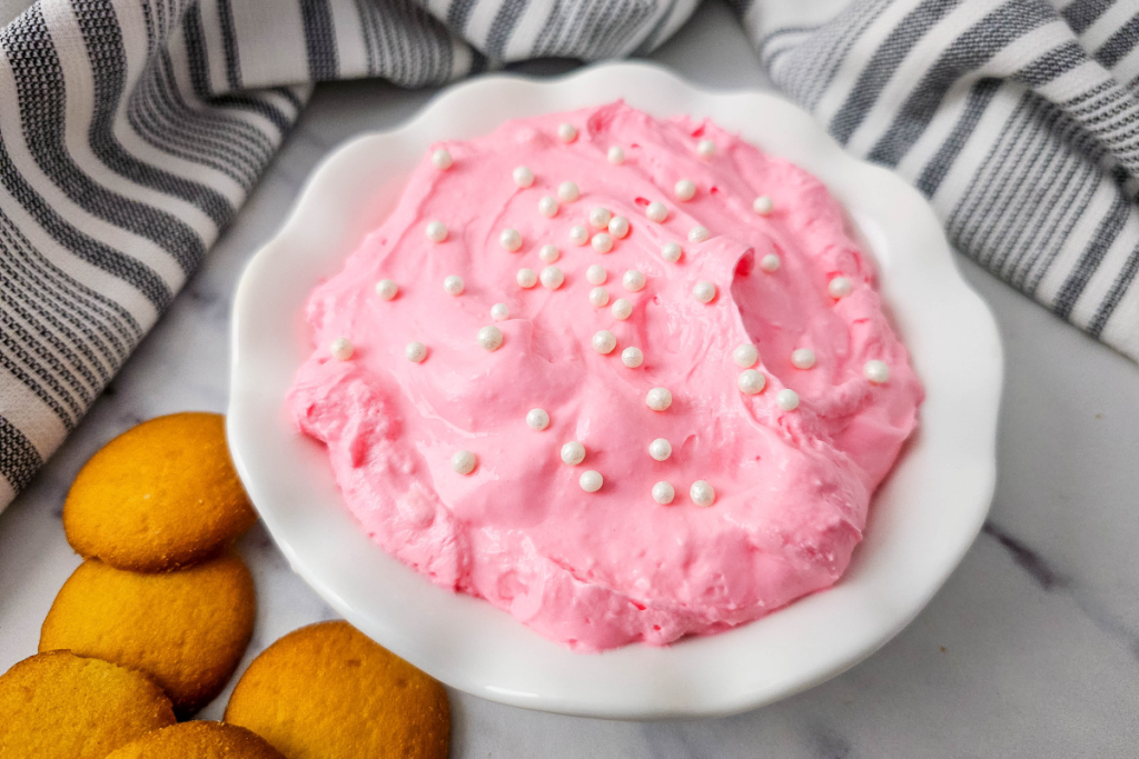 The finished Pink Fluff Dip served in a ruffled white bowl, garnished with white pearl sprinkles, next to a stack of golden vanilla wafer cookies.