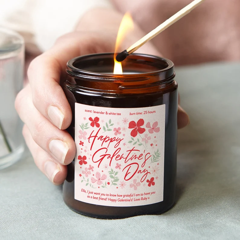 A hand lighting a small amber candle labeled "Happy Galentine's Day" with a personalized message, a cozy party favor idea.