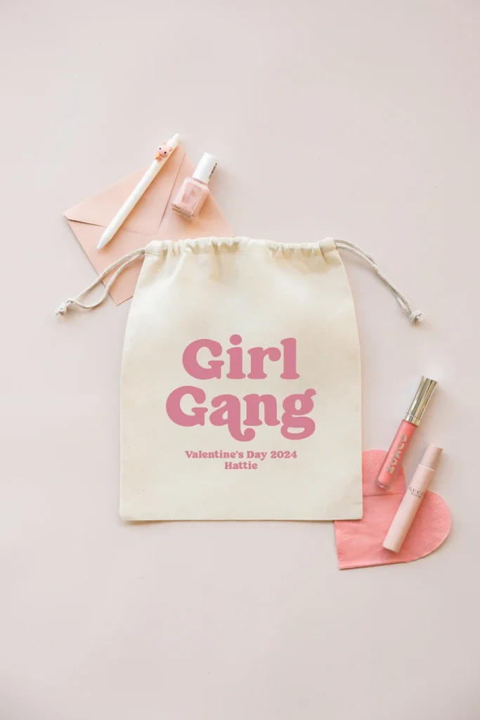 A cream drawstring bag with "Girl Gang" printed in pink, surrounded by makeup and nail polish, ideal for Galentine's Day Party Favors.