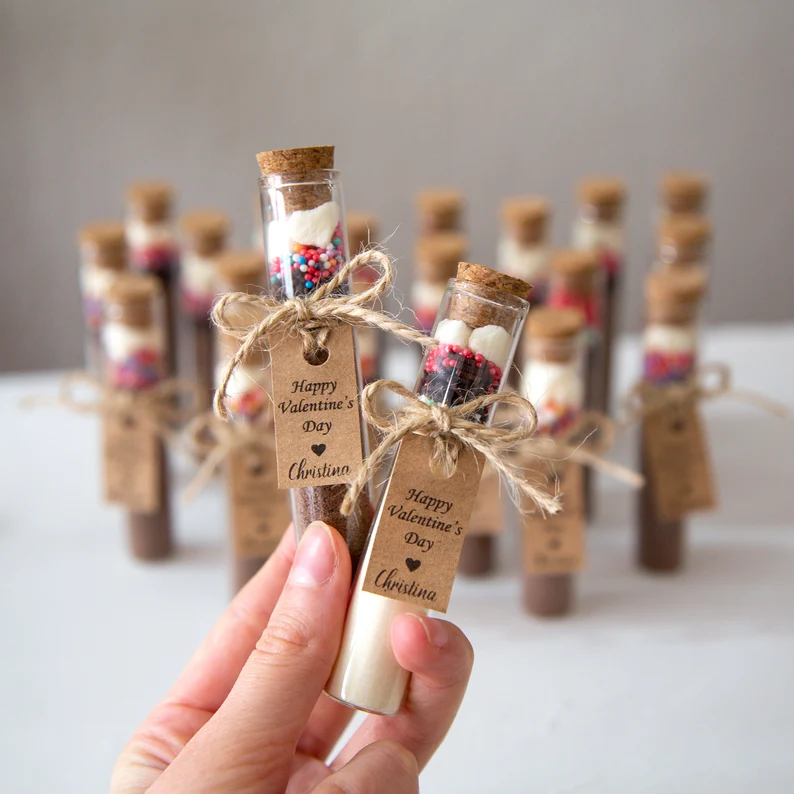 Two corked test tubes containing layers of white and dark hot chocolate mix with colorful sprinkles, finished with a personalized "Christina" tag for Galentine's Day Party Favors.