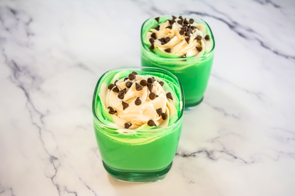 Two finished shots of the vibrant green Baileys Mint Cocktail are perfectly garnished with whipped cream and mini chocolate chips.