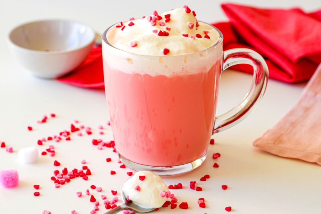A finished Red Velvet Hot Chocolate drink in a glass mug, topped with whipped cream and red heart sprinkles on a white table.