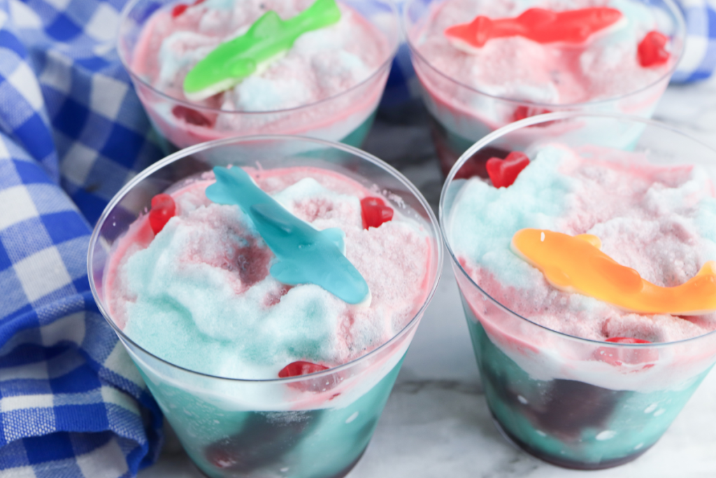 Four finished servings of blue and red Shark Attack Slush drinks, each garnished with a colorful gummy shark candy.