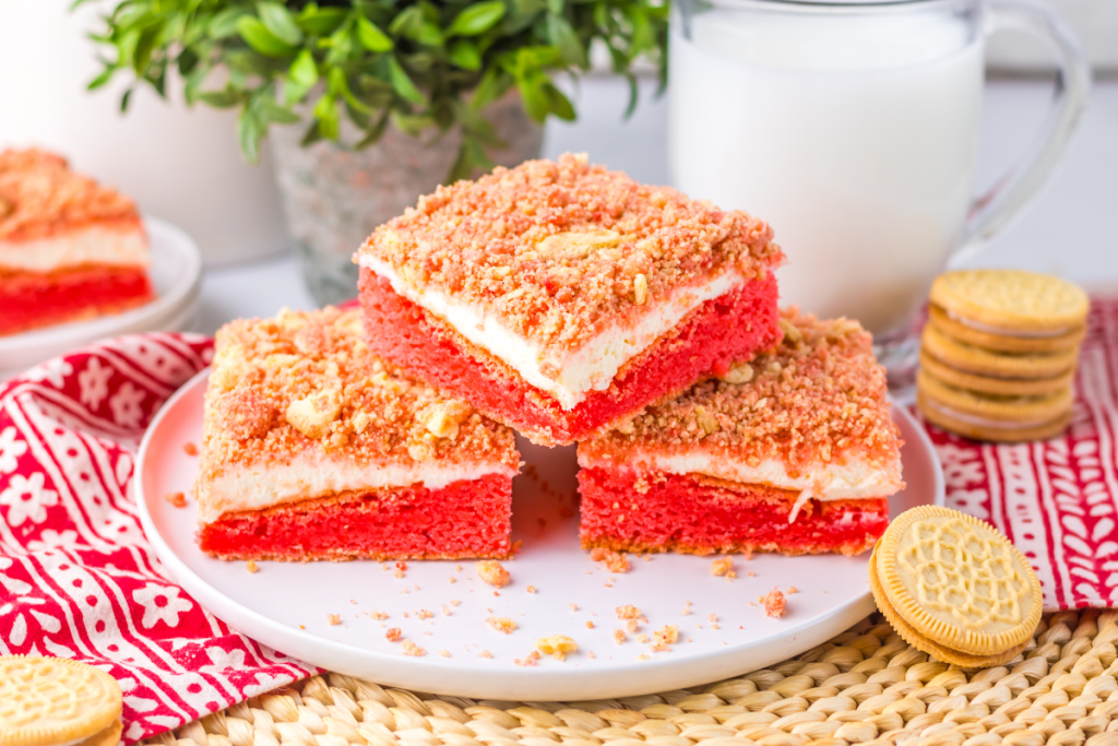 Three finished square slices of layered Strawberry Cheesecake Brownies stacked on a white plate, featuring a red base, white cheesecake layer, and a crumb topping.