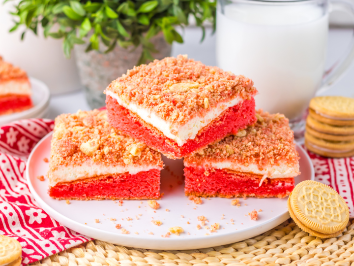 Three finished square slices of layered Strawberry Cheesecake Brownies stacked on a white plate, featuring a red base, white cheesecake layer, and a crumb topping.