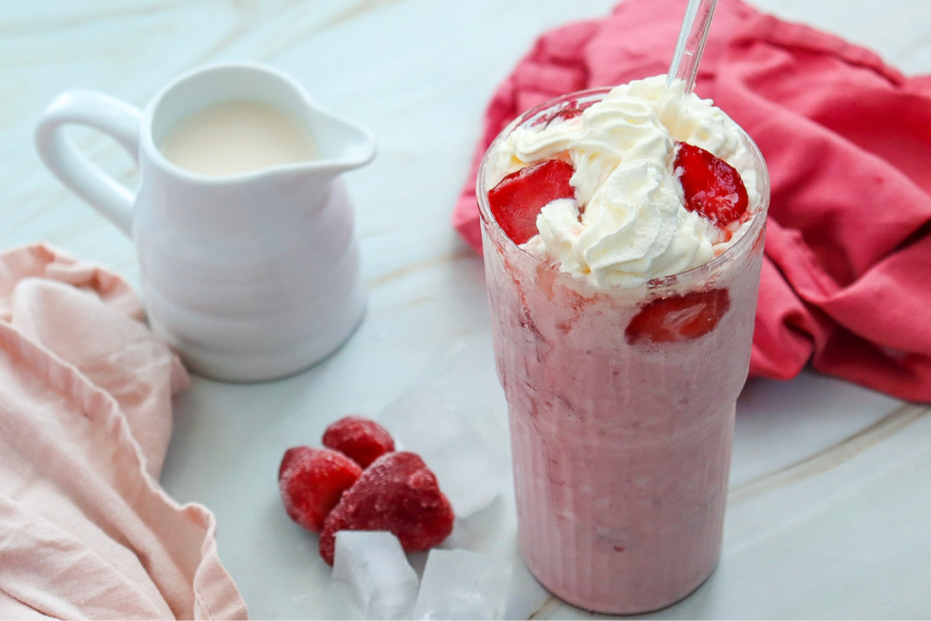 A tall glass of blended Strawberry Cheesecake Frappuccino topped with whipped cream and fresh strawberry pieces on a marble surface.